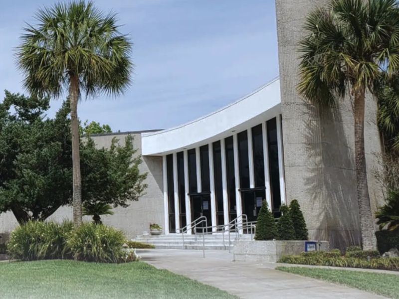 front church steps A modern building with a curved design, surrounded by palm trees and greenery, featuring steps leading to glass entrance doors.