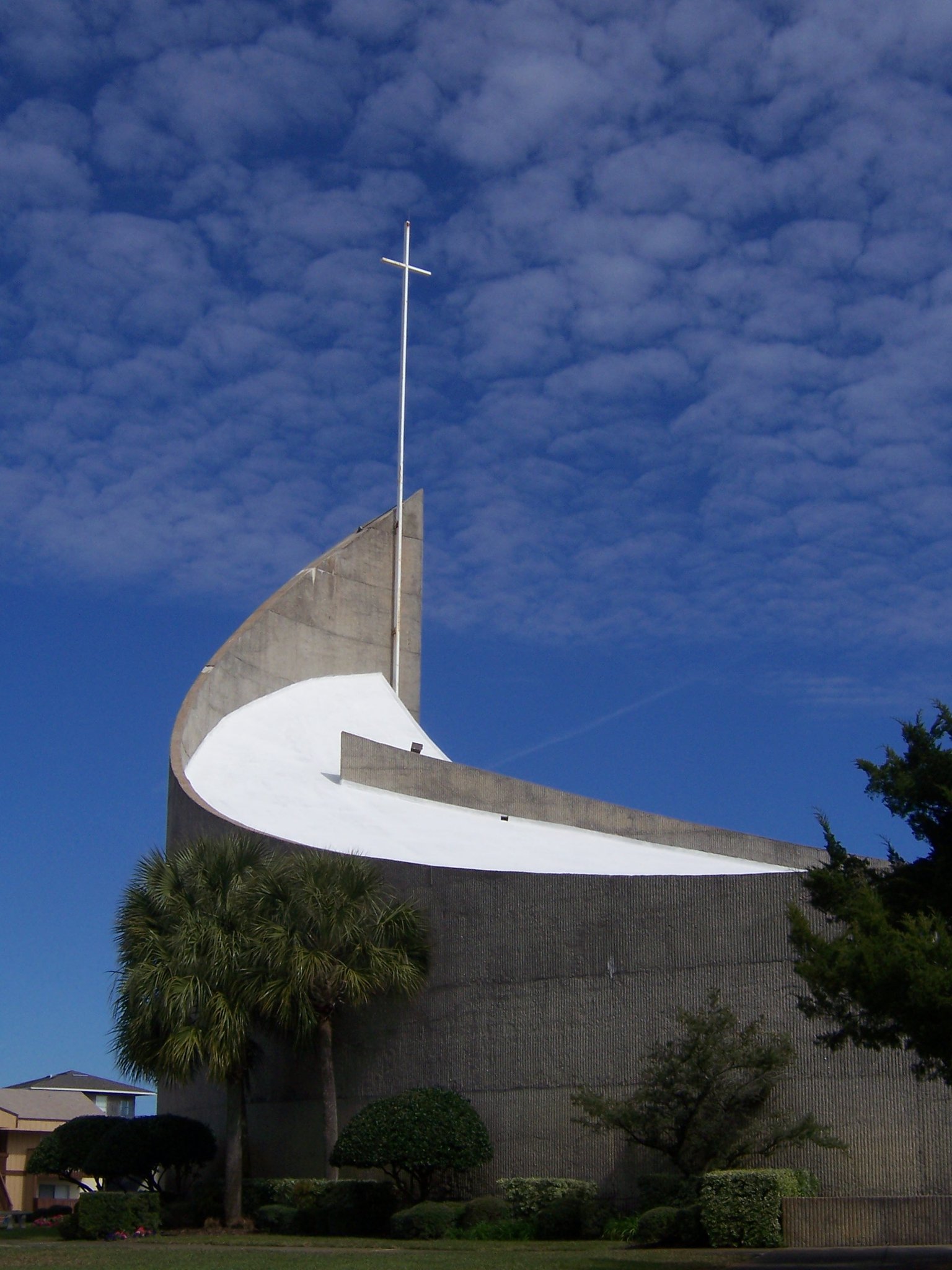 A modern church with a tall cross rises against a blue sky, surrounded by palm trees and lush greenery.
