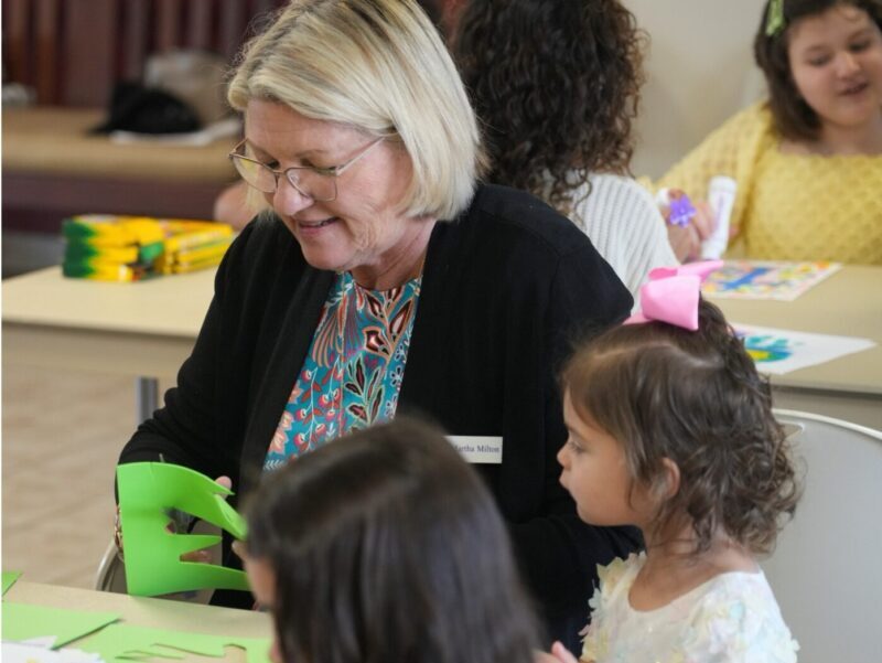 Picture13 A person and children engage in crafts at a table, cutting green paper. Smiling faces suggest a creative and educational activity.