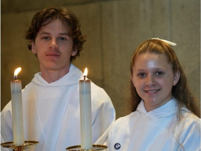 Two people in white robes hold tall lit candles, standing indoors, exuding a ceremonial atmosphere. They wear subtle expressions of calm focus.