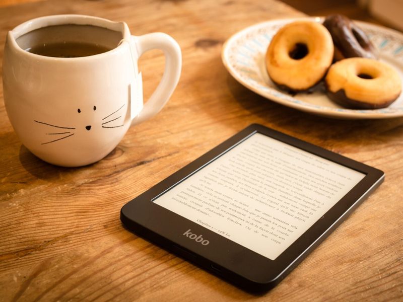 E-reader on a wooden table with a cat-themed mug of tea and a plate of donuts in the background.