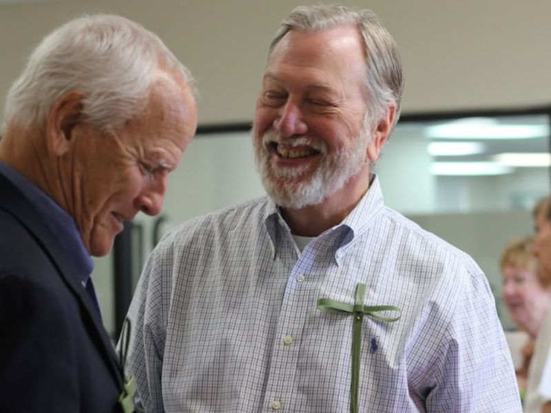 Two persons wearing shirt and jacket smiling in a room with a cross decoration. Background features blurred people and windows.